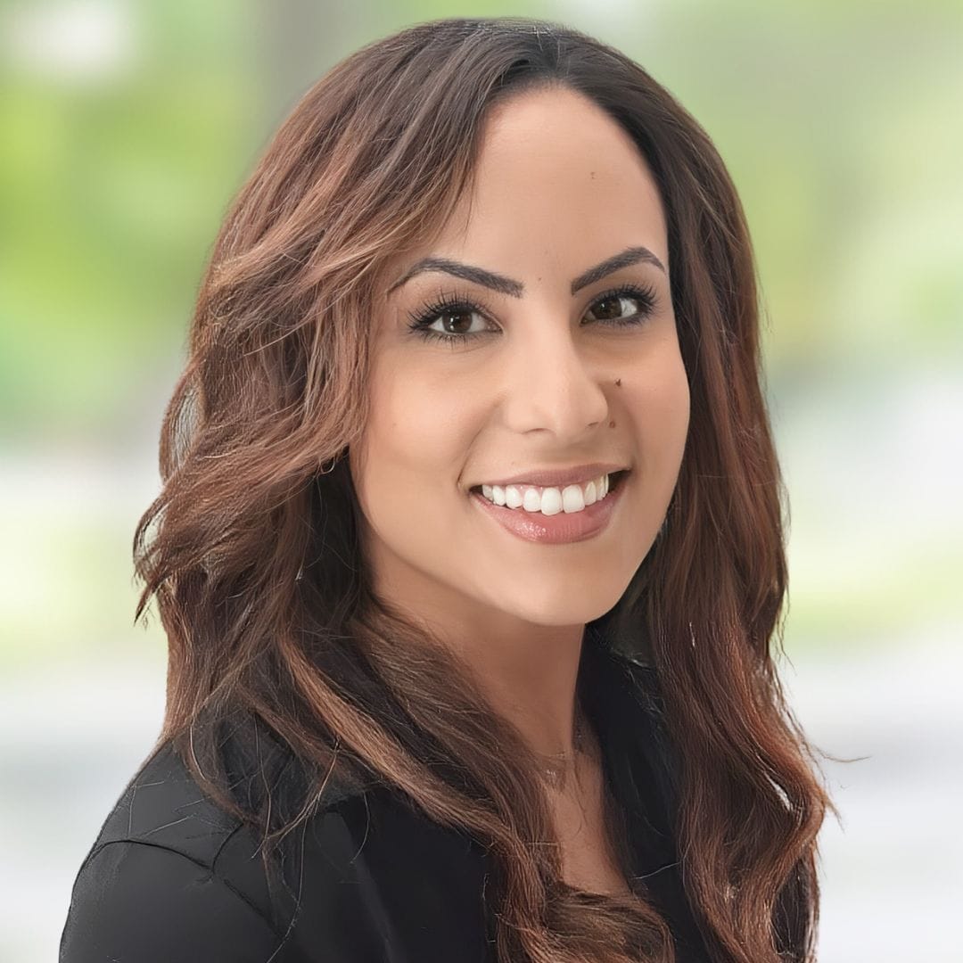 Cindy Derequito, a Psychiatric Mental Health Nurse Practitioner with long wavy brown hair, smiles at the camera in a black top. The softly blurred green and white background suggests an outdoor or natural setting.
