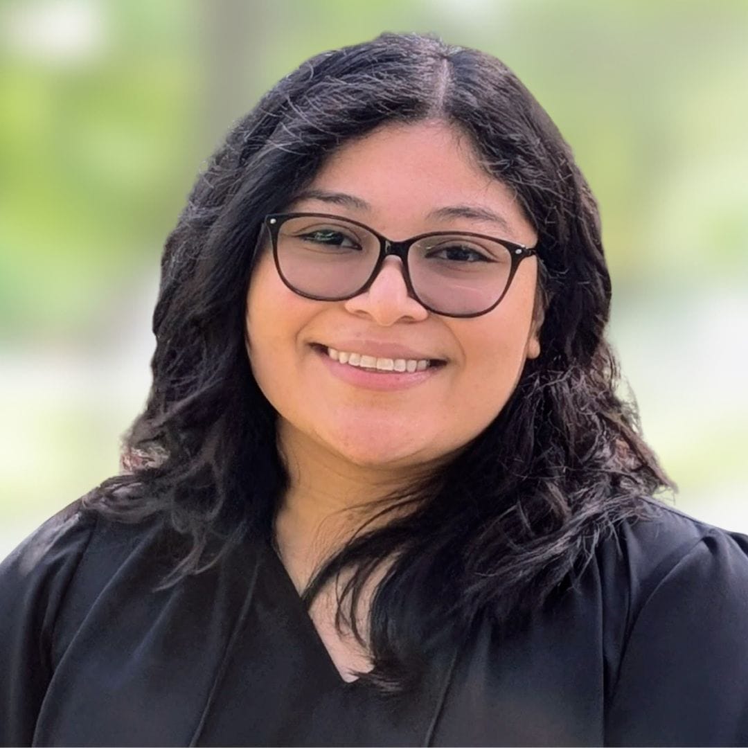 A young woman with wavy black hair and glasses smiles at the camera. She is wearing a black top and standing in front of a blurred, green outdoor background.