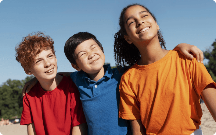 Three kids sit close together outdoors, smiling and squinting in the bright sunlight. One child has an arm around the others. They wear red, blue, and orange shirts, with trees and blue sky in the background.