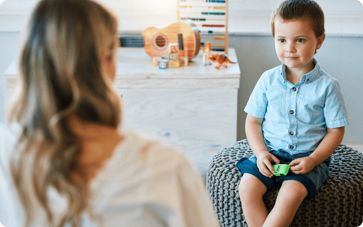 A young boy sits on a pouf holding a toy, facing an adult woman in a light room with toys and a guitar in the background.