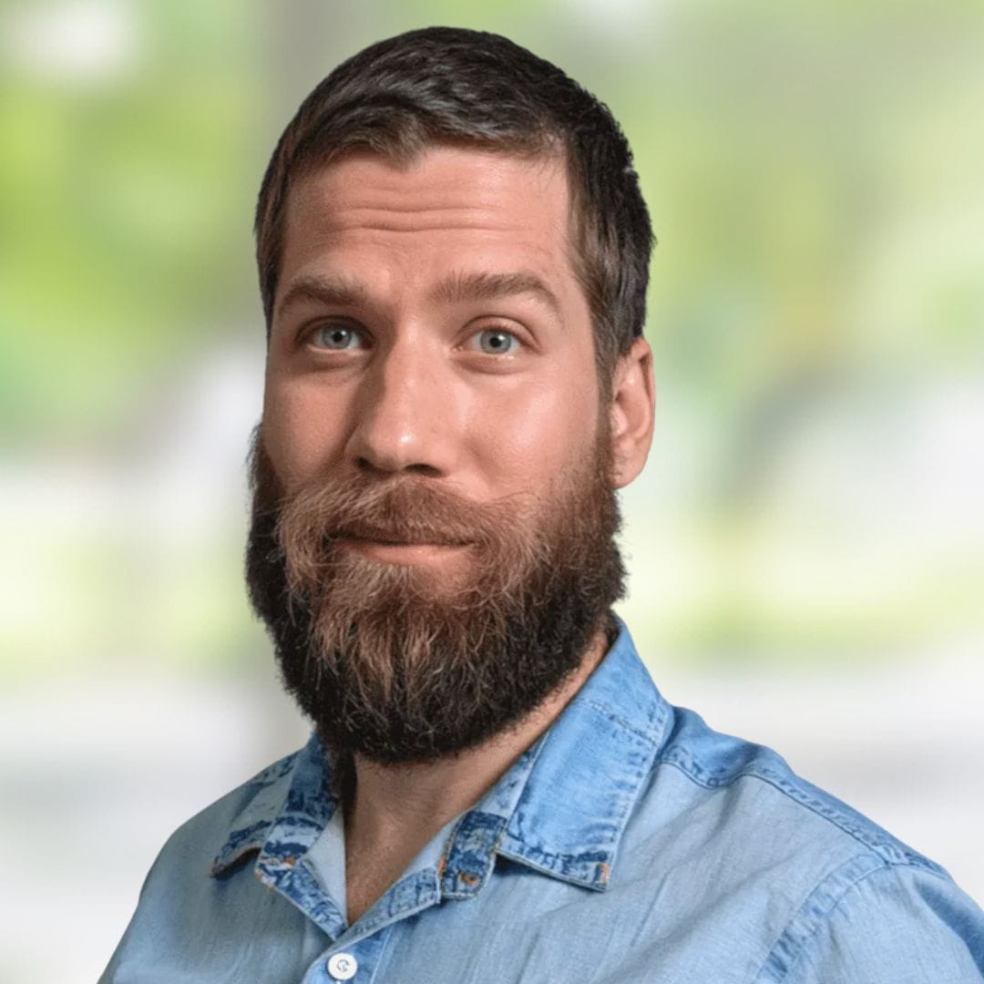 A man with a full beard and short brown hair wearing a light blue denim shirt stands in front of a blurred green and white background, smiling slightly.