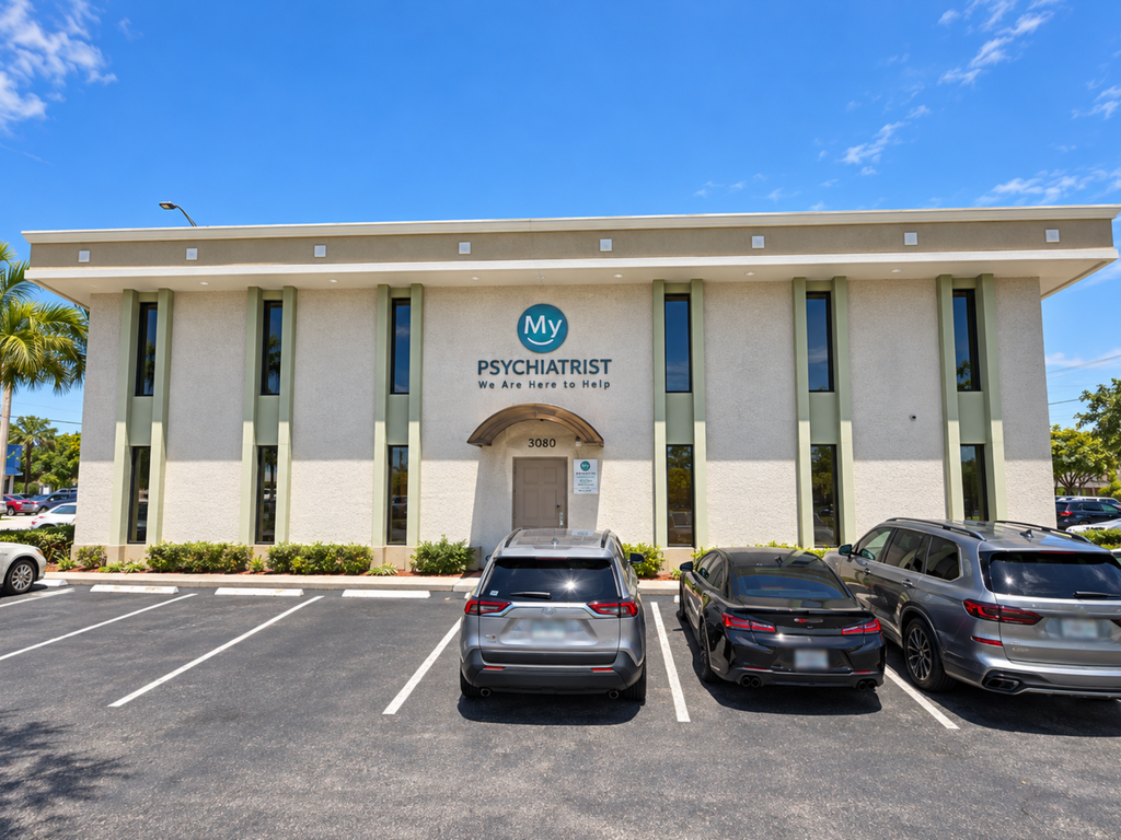 A beige two-story building with My Psychiatrist Oakland Park and We Are Here to Help on the front. Three cars are parked in front, with a clear blue sky above.