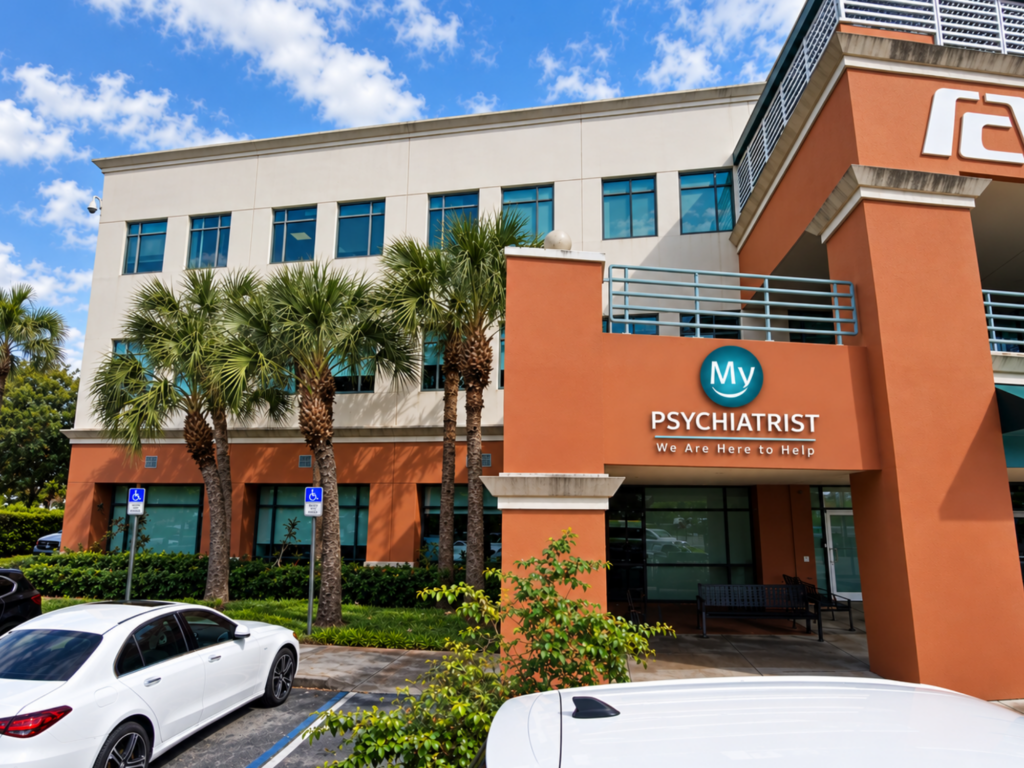 A modern building with palm trees in front, displaying a sign that reads My Psychiatrist - We Are Here to Help, offers mental health treatment Sheridan residents can trust. Several cars are parked in the lot, and the sky is partly cloudy.