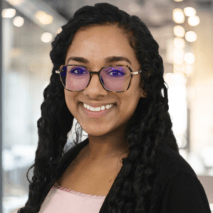 A woman with long curly hair, glasses, and a black sweater smiles at the camera in a modern office setting with blurred lights in the background.
