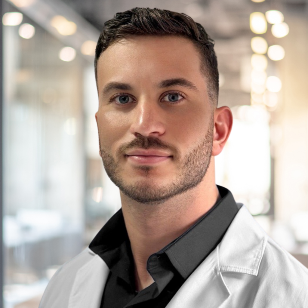 A man with short dark hair and a beard wearing a white medical coat over a black collared shirt.