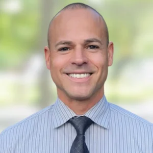 A smiling man with a shaved head wearing a light gray striped shirt and a dark gray tie, standing in front of a blurred green and white outdoor background.