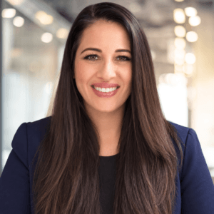 A woman with long brown hair wearing a navy blazer and black top smiles in a bright, modern office with glass walls and blurred lights in the background.