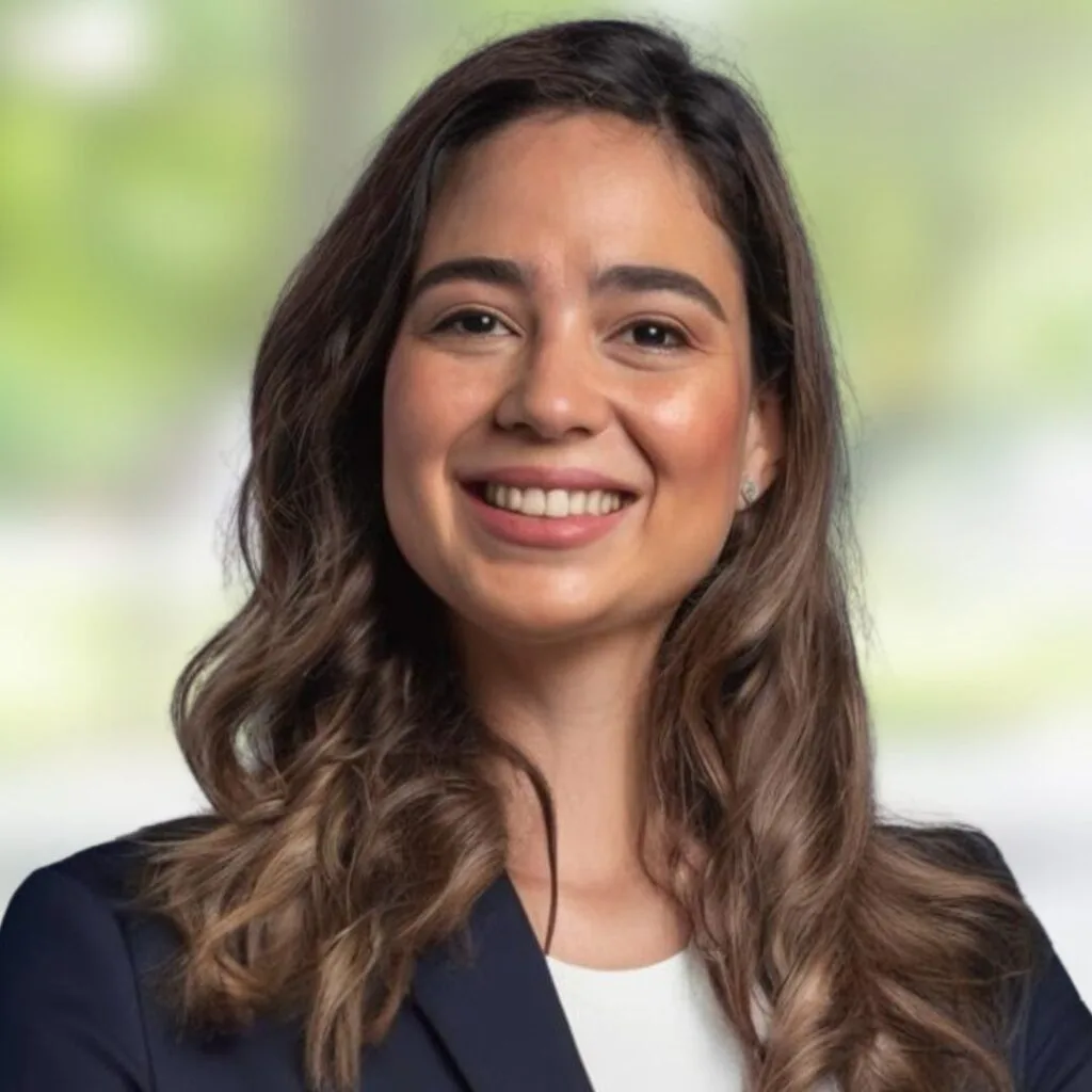 A woman with long wavy brown hair, wearing a navy blazer and white top, smiles confidently at the camera. The background is softly blurred with green and light colors.