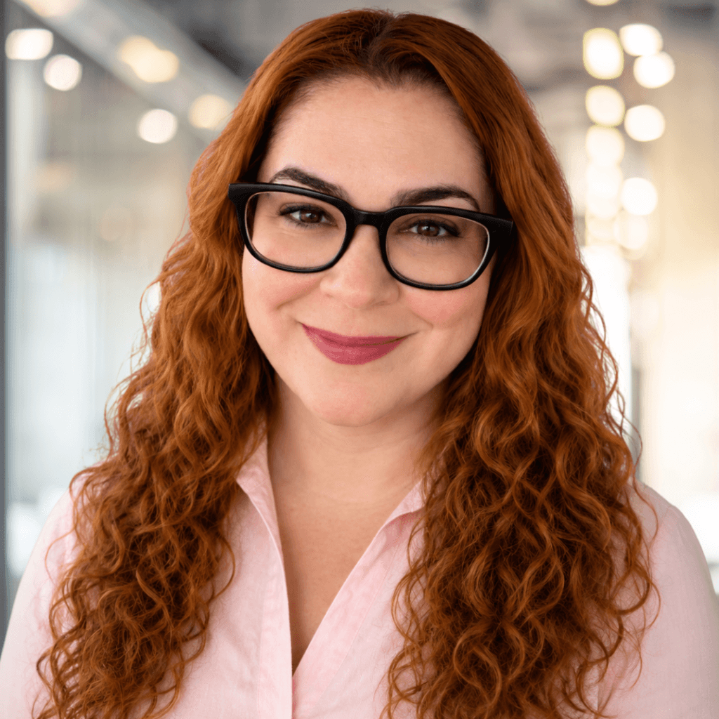 A woman with long curly red hair and glasses smiles, standing indoors in a bright, modern office.