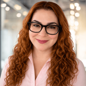 A woman with long curly red hair and glasses smiles, standing indoors in a bright, modern office.