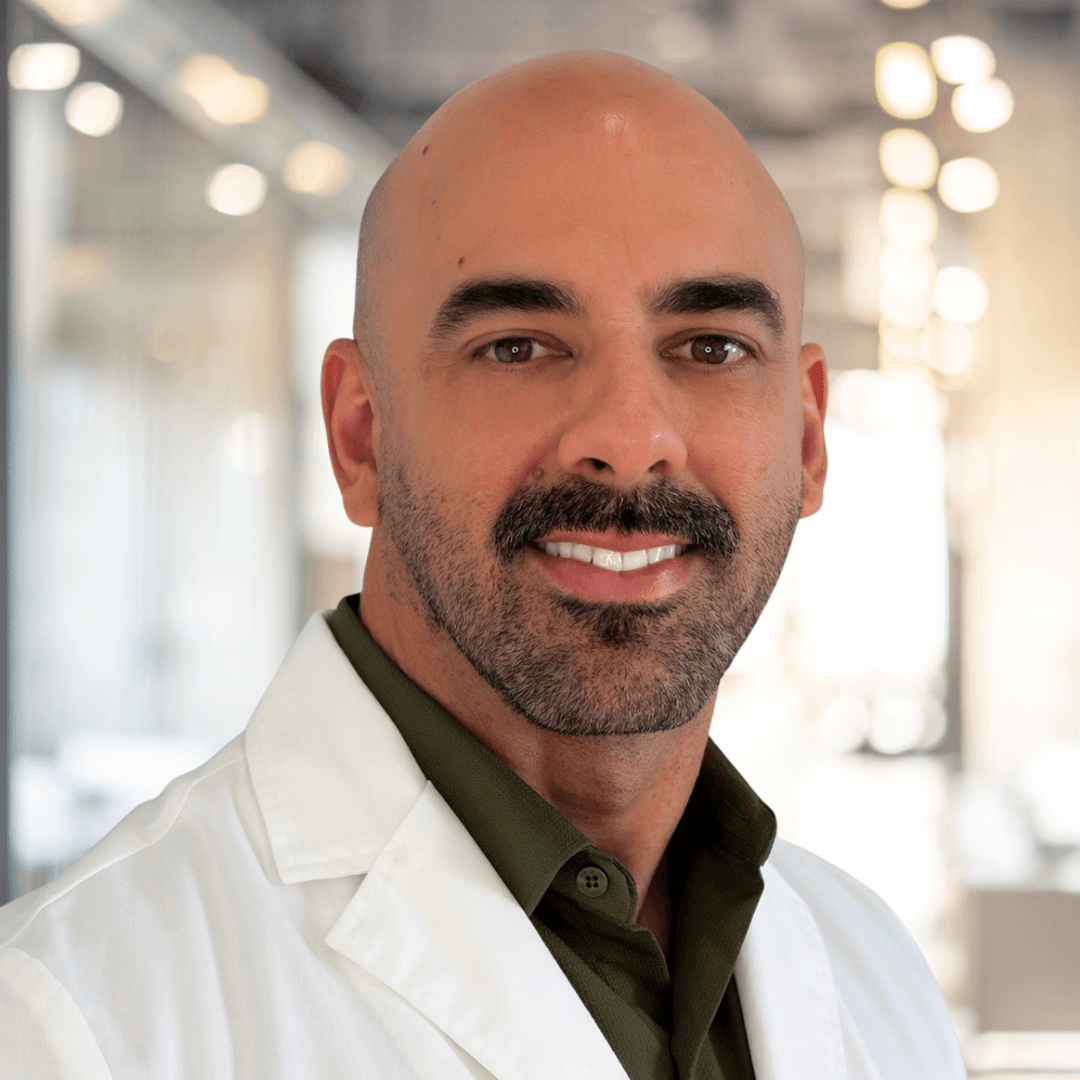A man with a bald head, beard, and mustache wearing a white lab coat and dark green shirt smiles in a bright, modern office or medical environment.