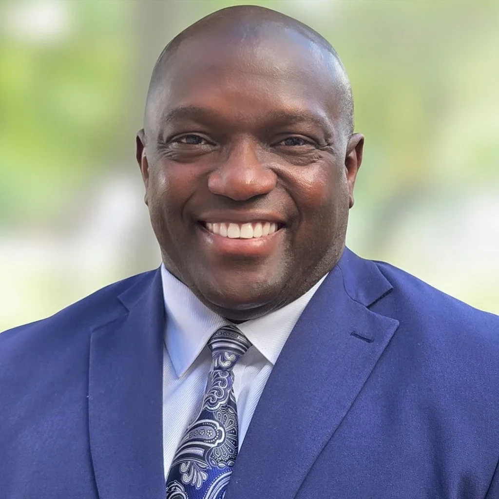 A man wearing a blue suit, white shirt, and patterned tie smiles at the camera against a blurred outdoor background.
