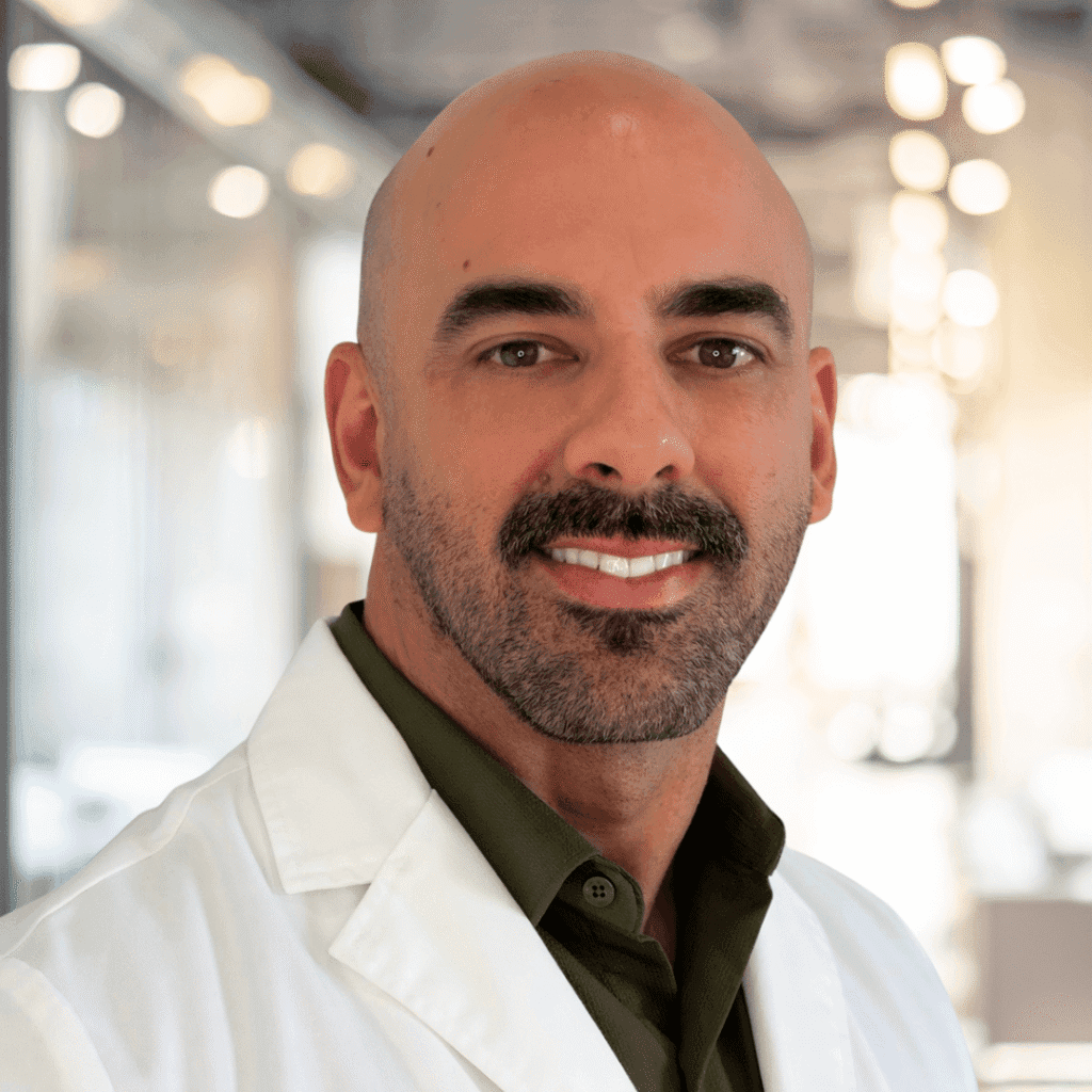 A man with a bald head, trimmed beard, and mustache is wearing a white lab coat and dark shirt, smiling in a bright, modern indoor setting.
