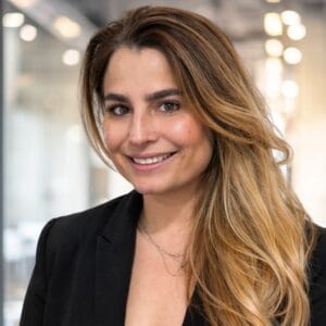 A woman with long, wavy light brown hair and a black blazer smiles at the camera in a modern, softly-lit office setting.