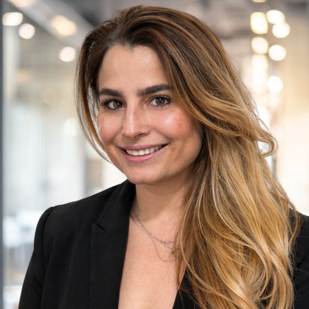 A woman with long, wavy light brown hair and a black blazer smiles at the camera in a modern, softly-lit office setting.