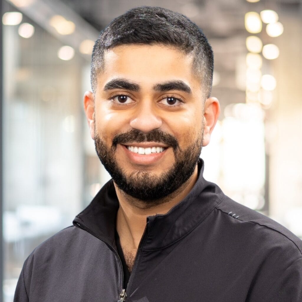A man with short dark hair and a beard smiles at the camera, wearing a black zip-up jacket in a modern, brightly lit office setting.