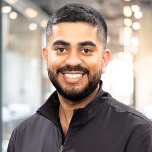 A man with short dark hair and a beard smiles at the camera, wearing a black zip-up jacket in a modern, brightly lit office setting.