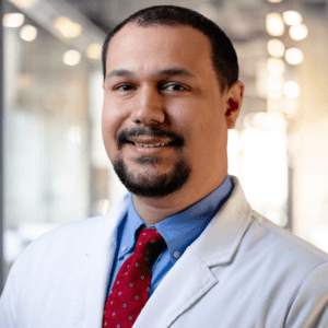 A man wearing a white lab coat, blue shirt, and red tie is smiling in a brightly lit hallway with blurred lights and glass walls in the background.