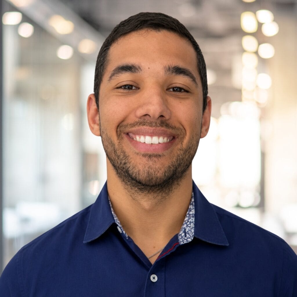 A young man with short dark hair and a beard smiles, wearing a dark blue collared shirt. He stands in a bright, modern office hallway with blurred lights in the background.