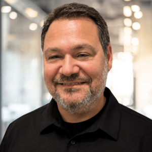 A man with short dark hair and a salt-and-pepper beard smiles, wearing a black collared shirt. He stands indoors with glass walls and blurred lights in the background.