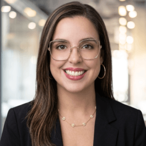 A woman with long brown hair, wearing glasses, a black blazer, gold hoop earrings, and a necklace, smiles in a modern office setting with blurred lights in the background.