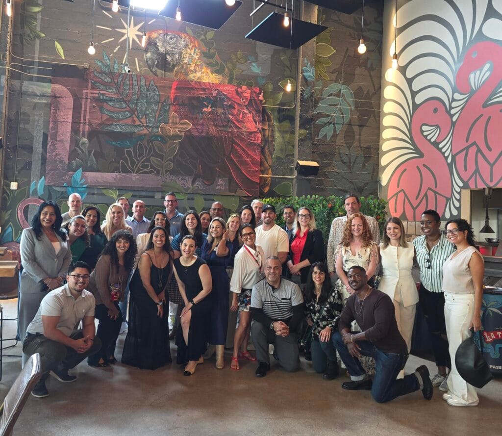 A group of about 30 people pose and smile inside a stylish space with colorful wall art featuring a jaguar, leaves, and two pink flamingos. There are pendant lights and a plant in the background.