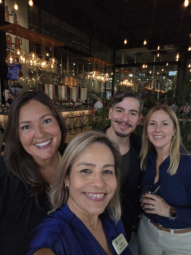 Four adults standing close together and smiling at the camera in a modern, warmly-lit bar or restaurant with hanging lights and a reflective ceiling behind them.