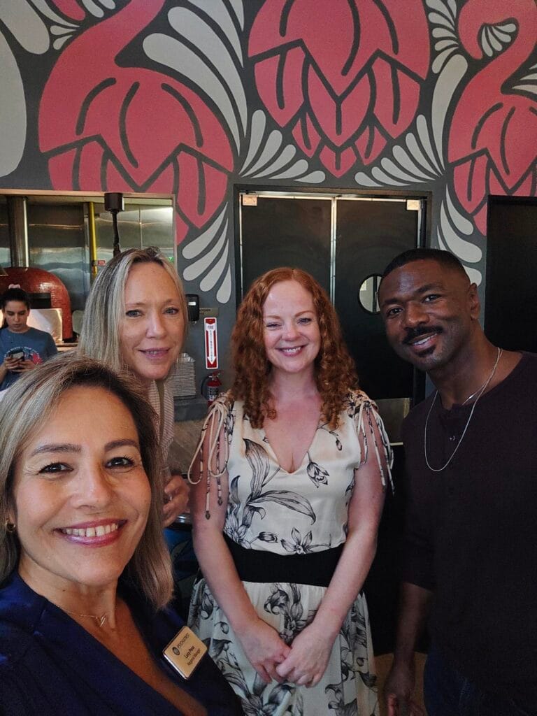 Four people smiling and posing for a photo indoors, with a wall featuring large pink and gray floral designs in the background. One person is taking the selfie.