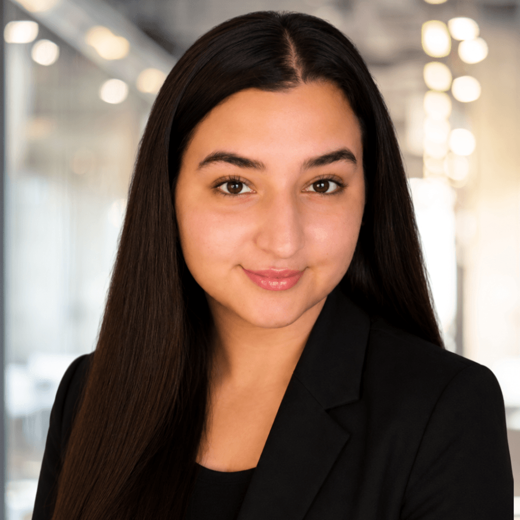 A woman with long, dark hair and a black blazer smiles softly in a modern, well-lit office setting with blurred lights in the background.