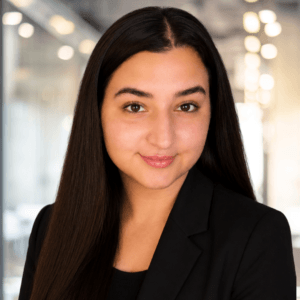 A woman with long, dark hair and a black blazer smiles softly in a modern, well-lit office setting with blurred lights in the background.