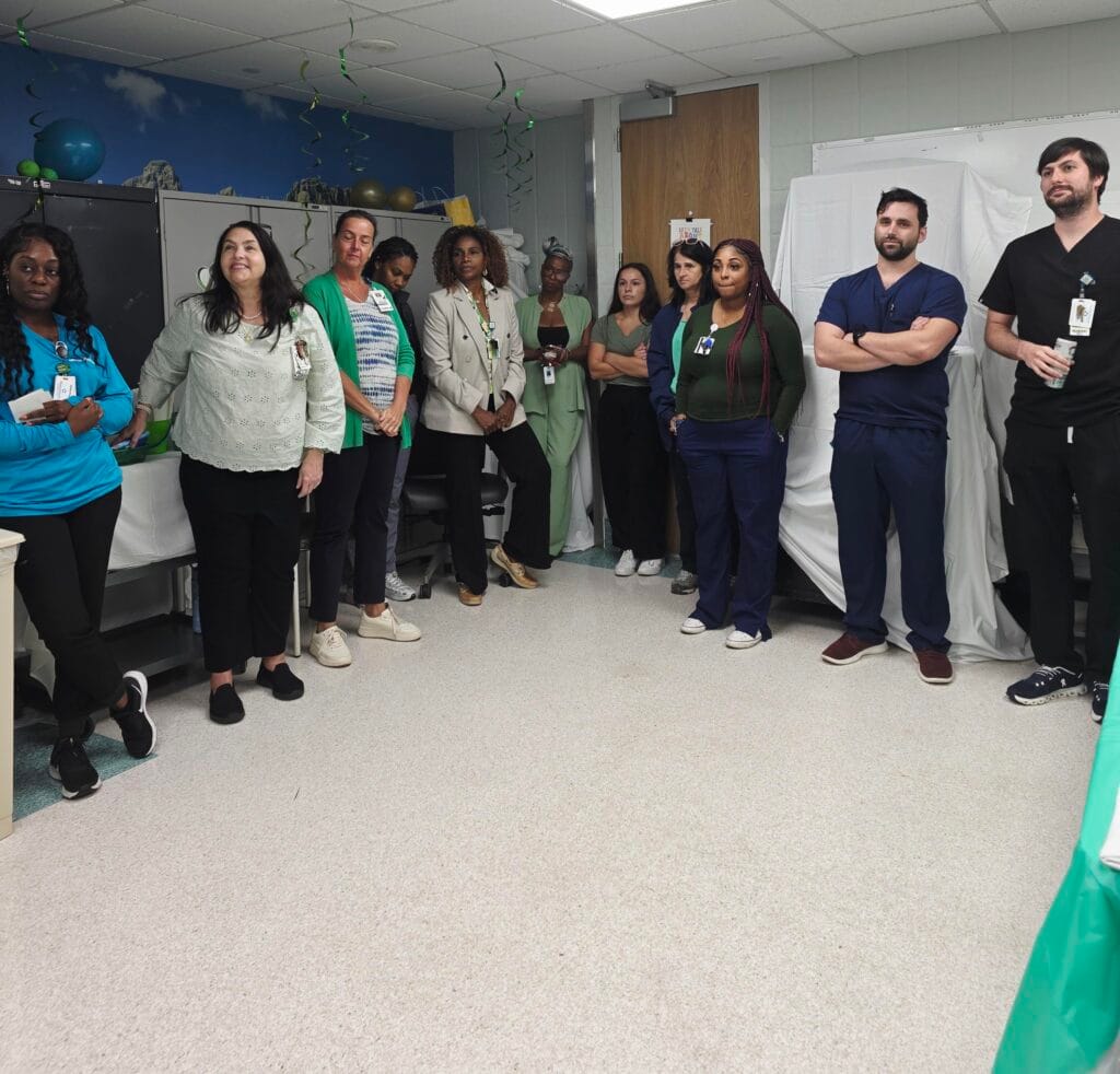 A group of healthcare professionals stands in a hospital room, listening attentively. Some wear scrubs while others are in business or casual attire. Decorations and medical equipment are visible in the background.