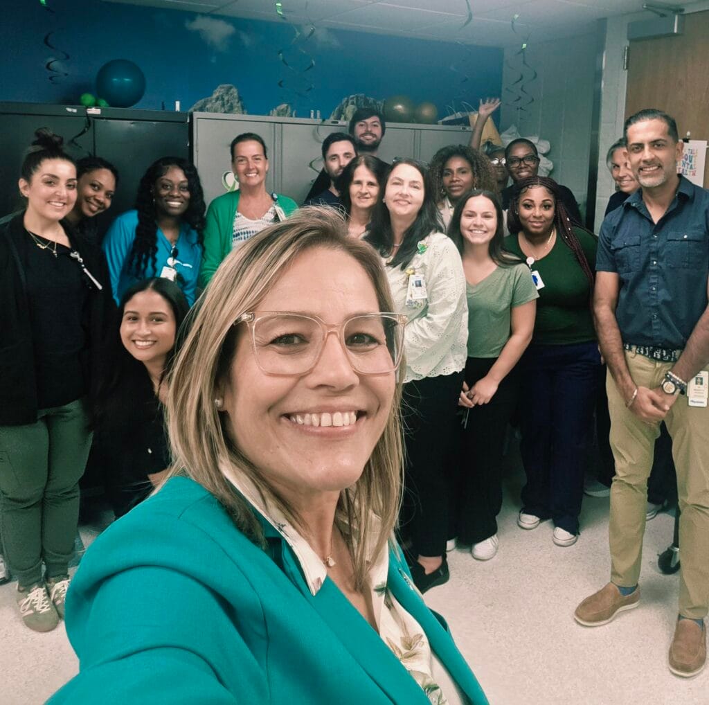 A group of people pose together for a selfie in an indoor setting, smiling at the camera. Decorations are visible on the ceiling, and most people are wearing business casual clothing.