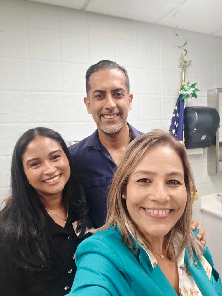 Three adults smiling for a selfie in an office with white walls, an American flag, a paper towel dispenser, and a plant visible in the background.