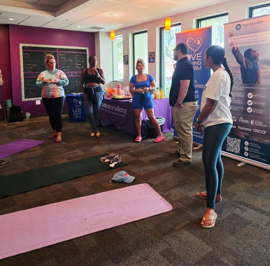 A group of people stand and chat in a room with yoga mats, wellness banners, and snacks. The setting appears to be an indoor wellness or yoga event.