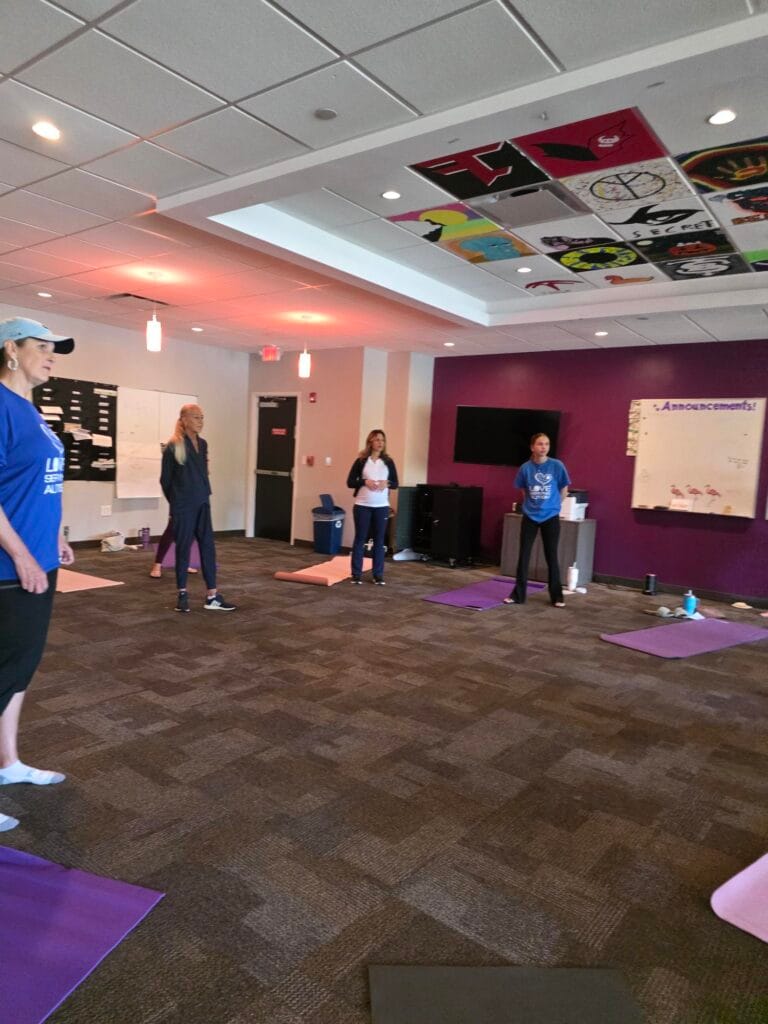 Four women stand on yoga mats in a spacious, brightly lit room with a purple accent wall, ceiling art, and announcement boards, preparing for a fitness or yoga class.