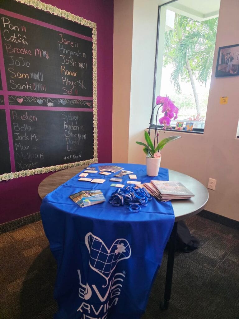 A round table with brochures, name tags, and blue lanyards is covered by a blue cloth with UVE ARVM. Behind it is a chalkboard with names and a window showing a tree and a blooming orchid on the sill.
