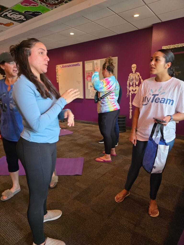 Four women stand in a brightly lit room with purple walls, exercising or talking. Two women in the foreground converse while others stretch. A skeleton decoration and fitness mats are visible in the background.