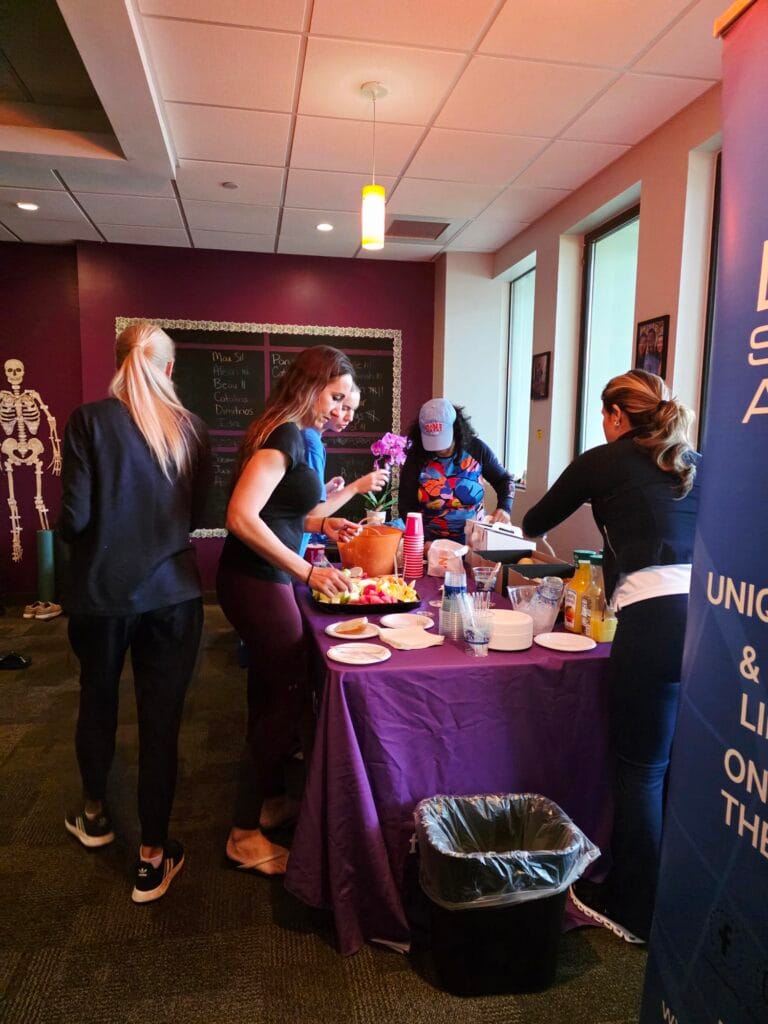Four women stand at a purple table with food and drinks in a room with a chalkboard and a skeleton model in the background. Plates, cups, and various food items are visible on the table.