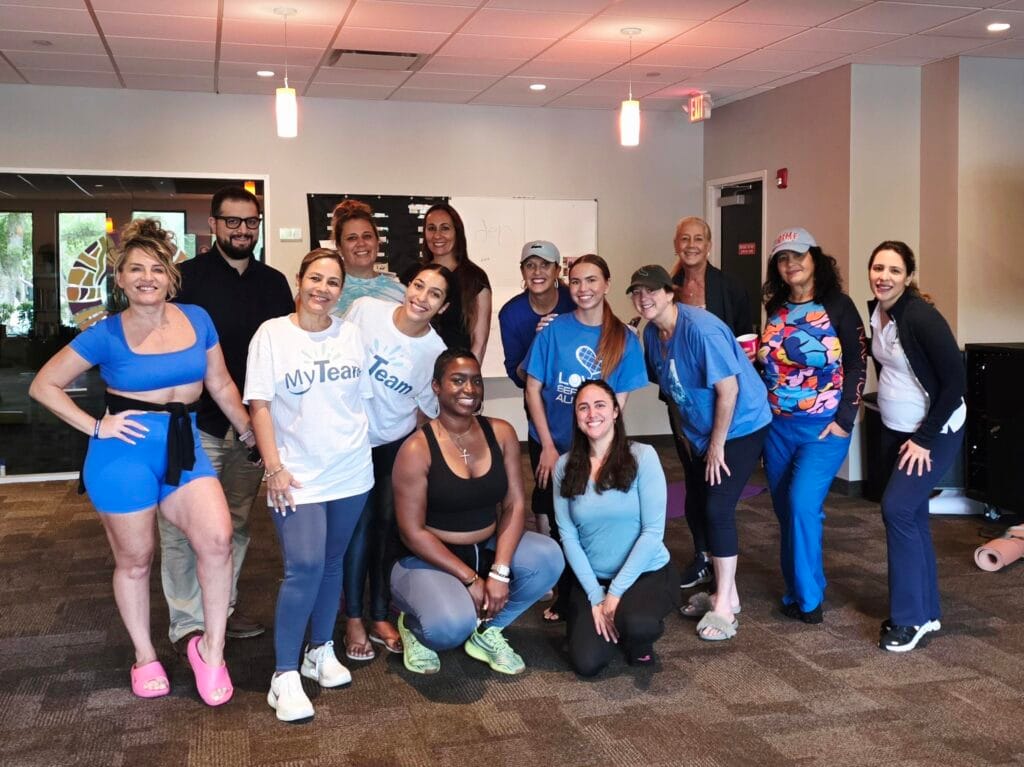 A group of people pose together indoors, some standing and some kneeling, all smiling. Most are wearing casual or athletic clothing in shades of blue and white. The setting appears to be a community or fitness center.