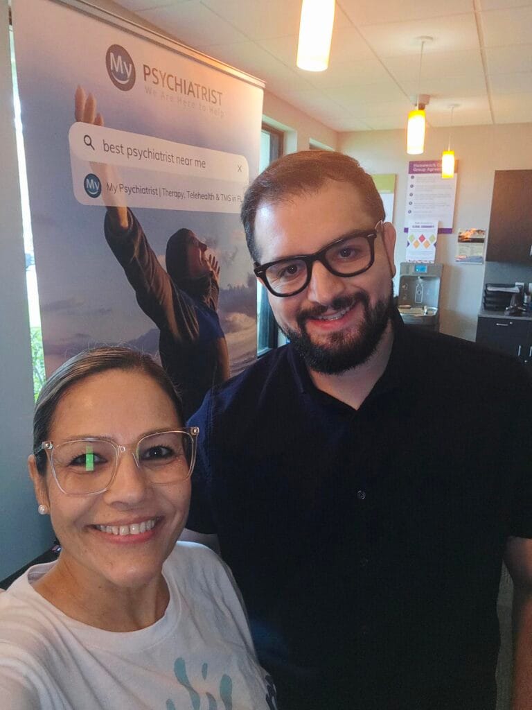 Two people smiling indoors, standing in front of a My Psychiatrist banner. The banner displays a search bar that reads best psychiatrist near me. Warm lighting and office furniture are visible in the background.