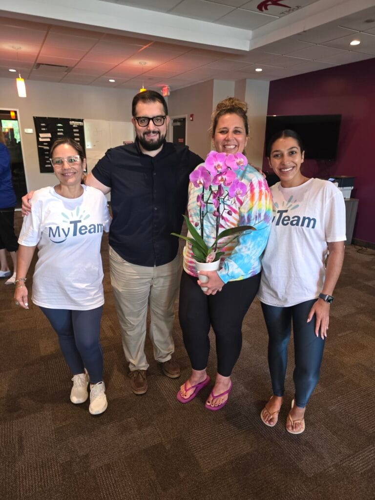 Four people stand indoors, two wearing MyTeam shirts. One person holds a potted orchid with purple flowers. They are smiling and standing on brown carpet with a purple accent wall and TV in the background.