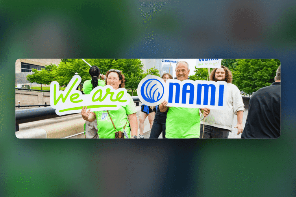 A group of people outdoors hold signs reading We are NAMI. They are smiling, wearing green shirts, and participating in what appears to be a community event or walk, likely as part of NAMIWalks Broward County.