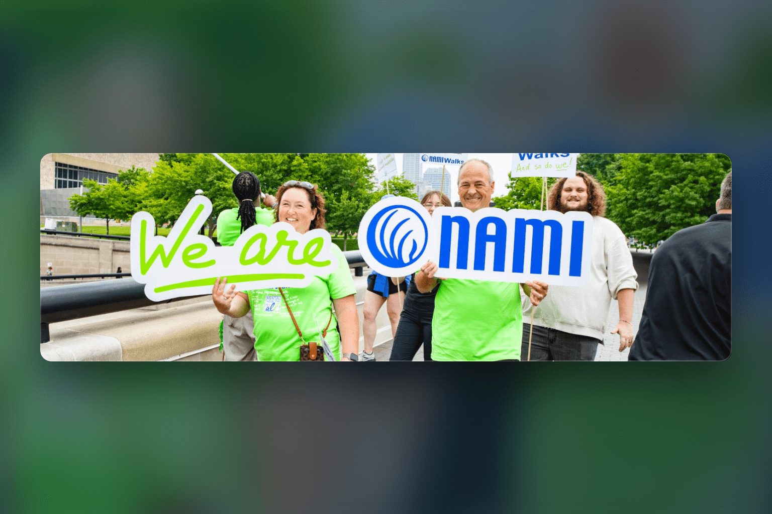 A group of people outdoors hold signs reading We are NAMI. They are smiling, wearing green shirts, and participating in what appears to be a community event or walk, likely as part of NAMIWalks Broward County.