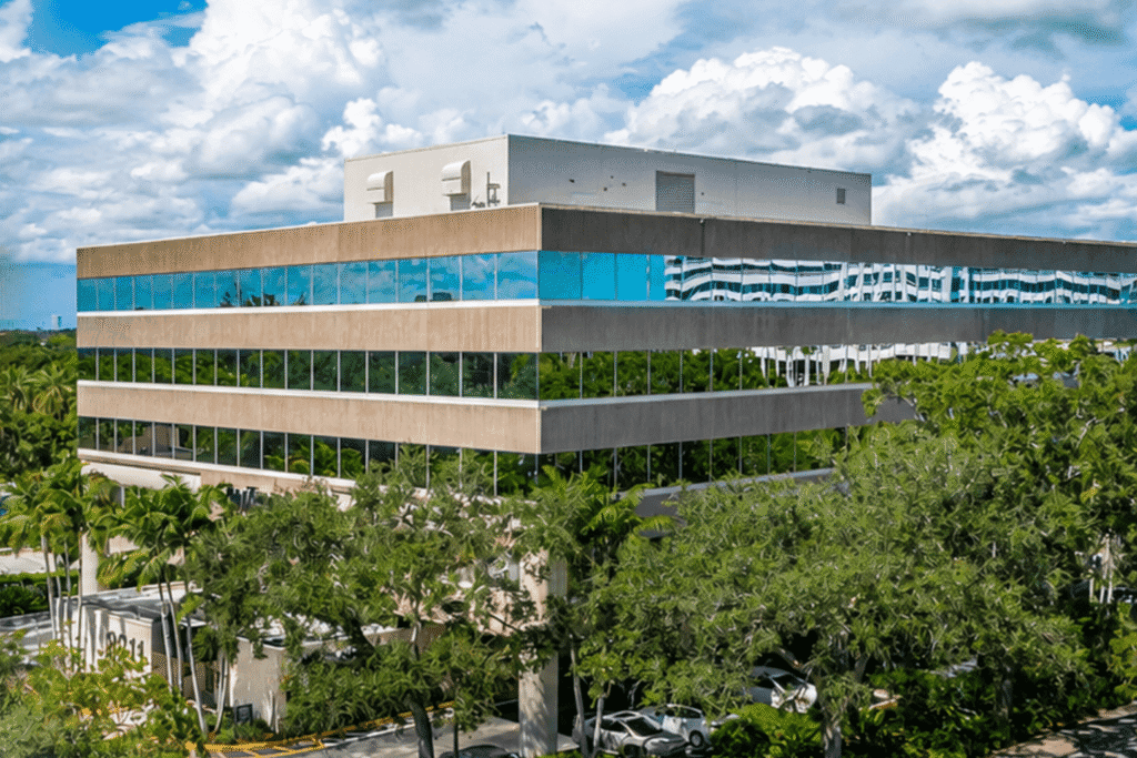 A multi-story office building with reflective blue windows, surrounded by green trees under a partly cloudy sky, now houses a new psychiatry office in Plantation. Several cars are parked near the entrance.