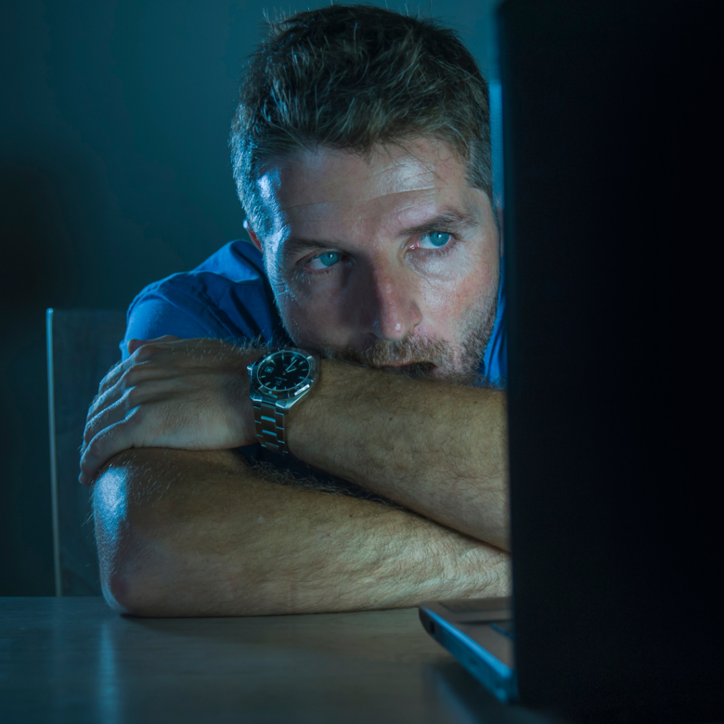 A man stares intently at his laptop screen in a dark room, searching for help with porn addiction.