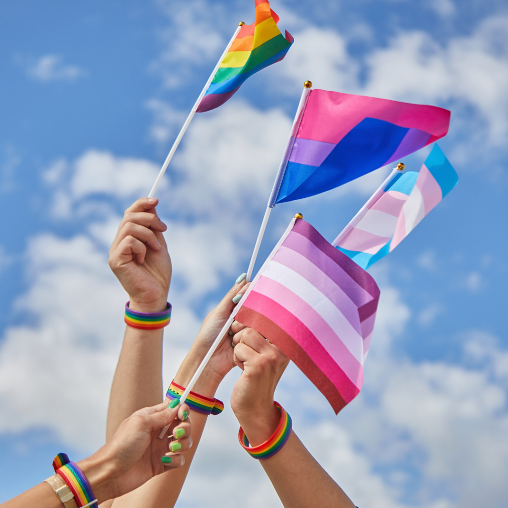 Hands held up against a blue sky, each wearing a rainbow wristband and waving a different pride flag in a symbol of LGBTQIA mental health support.
