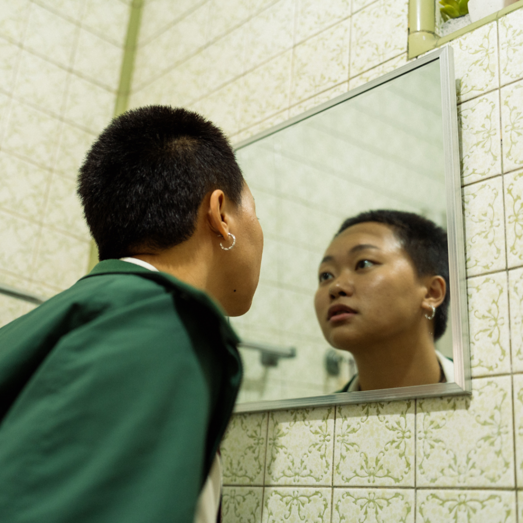 A person with short hair and hoop earrings looking at their reflection in a bathroom mirror, reflecting on their need for gender-affirming psychiatry.