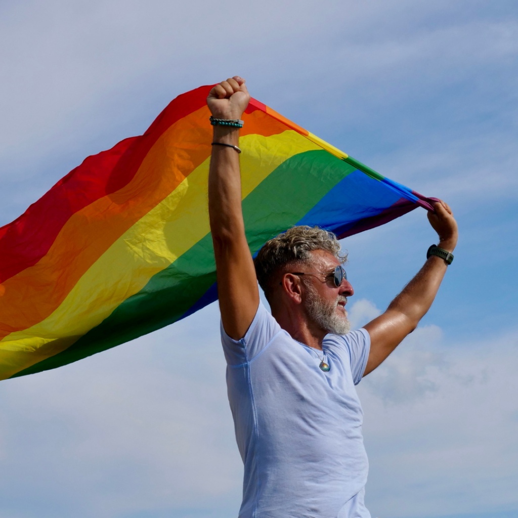 A man with a gray beard and sunglasses holds a rainbow flag high above his head against a blue sky, representing the supportive presence of a gender-affirming psychiatrist.