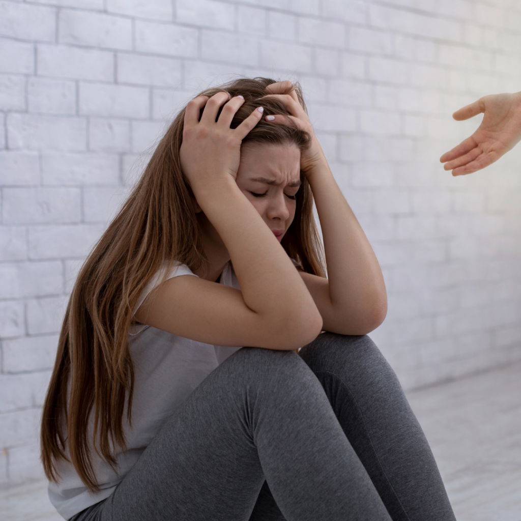 A distressed young woman sits on the floor holding her head in her hands while a ptsd psychiatrist reaches out to her with an open hand.