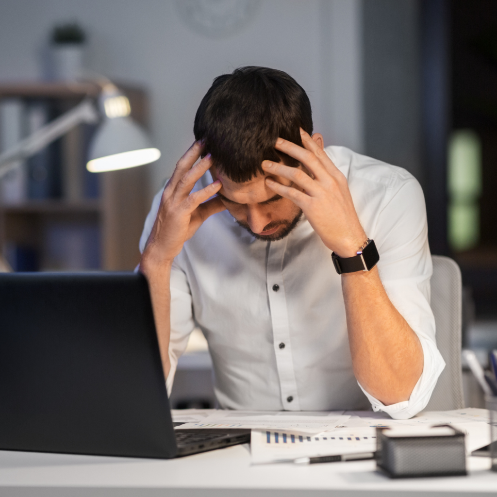 A man sitting at a desk with a laptop, holding his head in his hands with a stressed expression while looking for porn addiction help.